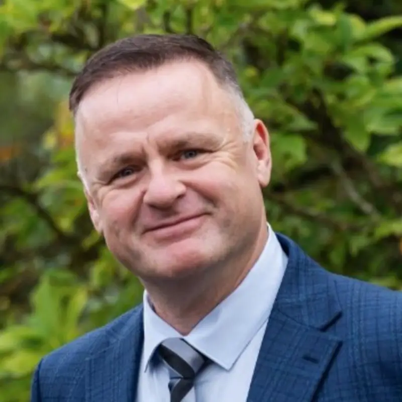 Image of Simon smiling to the camera. He is wearing a blue suit and shirt, with a striped tie.